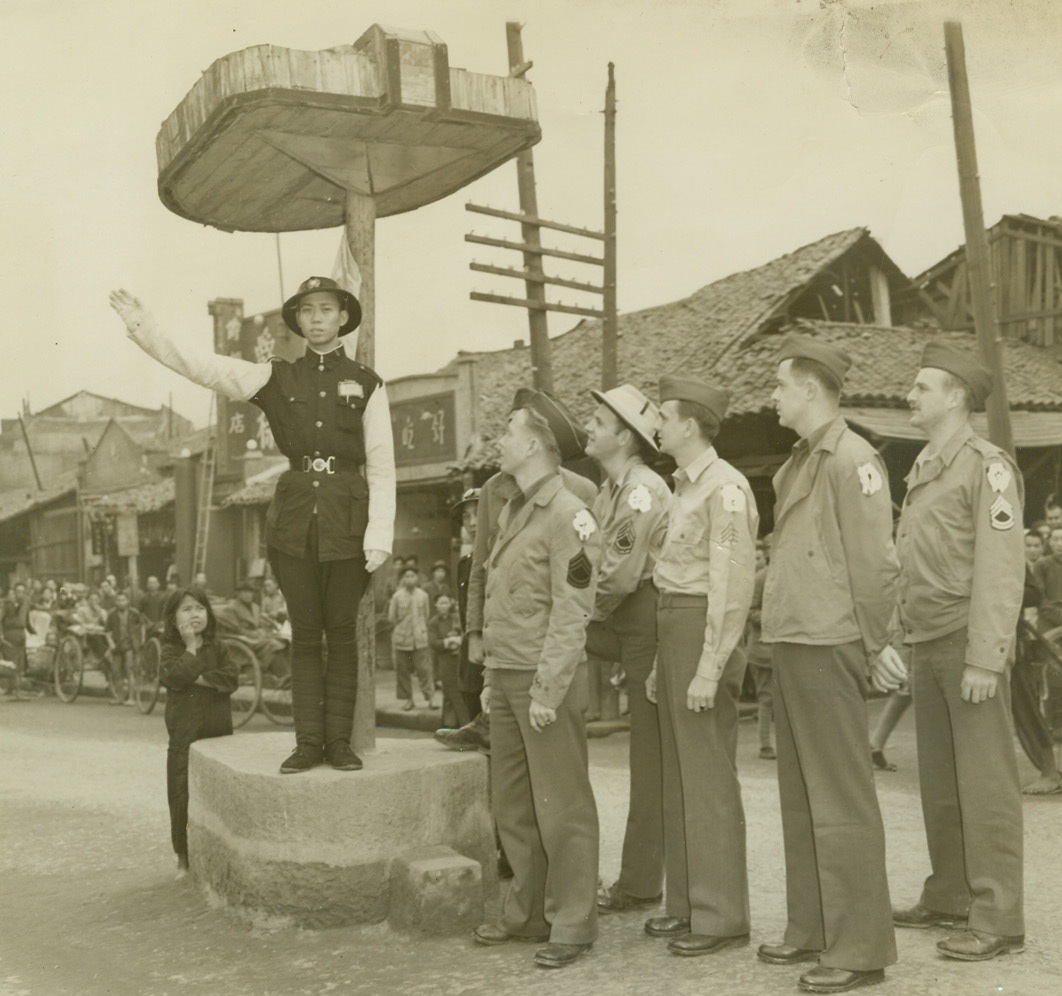 The Traffic is Terrific, 1/12/1943. Somewhere in China – American soldiers admire the efficiency and neat dress of a Chinese policeman who directs traffic at a “busy” intersection. Soldiers (left to right) are: Sgt. Ivan O. Stanberry, Pomona, Calif.; Claude J. Smith, N.C.; Sgt. Robert Wigglesworth, Wisc.; Sgt. Andrew Chemsakm PA.; and Sgt. Charles M. Janes, Calif. The little Chinese lady is as fascinated as the Americans. Credit: ACME;