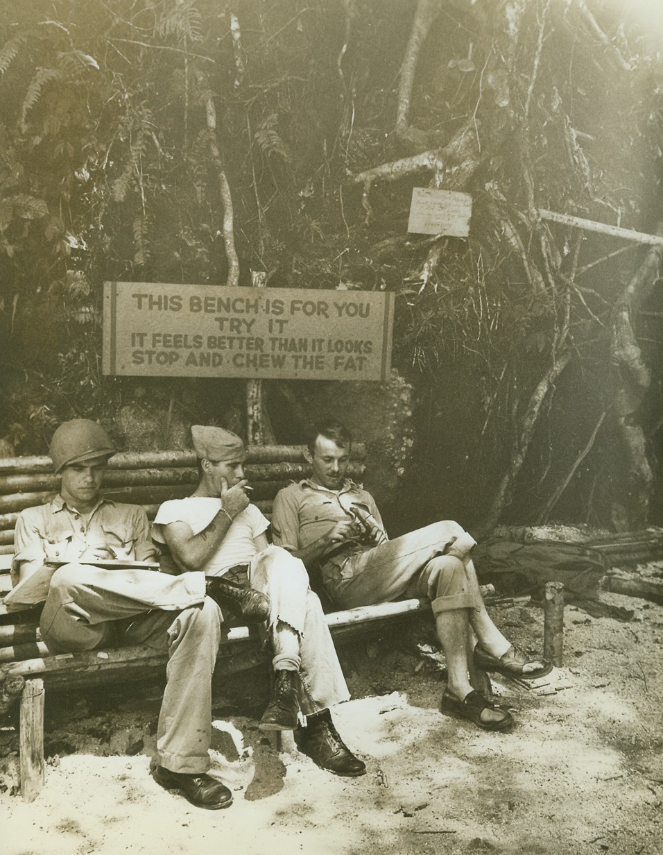 Bench and Sales Talk, 1/16/1943. Guadalcanal – These three Leathernecks were convinced by the sales talk tacked above the bench on Guadalcanal. One writes a letter home, another just smokes, and the third whittles – in  a pain of comfortable air-conditioned shoes.  They’re all enjoying a brief moment of relaxation in the tough tropical fighting zone. Credit line (US Marines Corps);