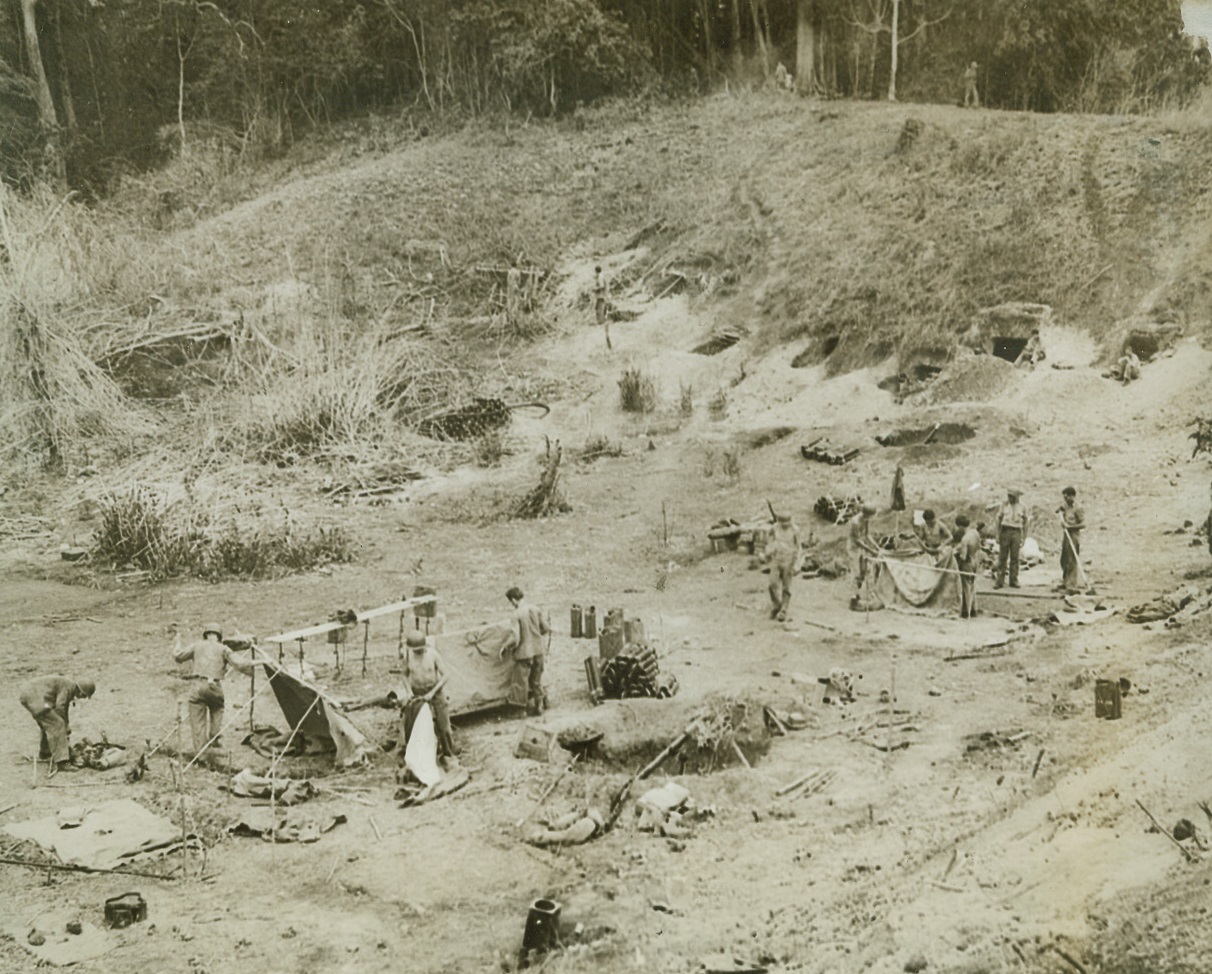 Guadalcanal “Cliff-Dwellers”, 1/16/1943. Guadalcanal, S.I. – Modern “cliff-dwellers”, these members of a U.S. Marine Corps mortar crew are living in a group of caves in a gulch on Guadalcanal.  The entrances to the caves, which the boys built themselves, can be seen at upper right of photo.  An armed sentry can also be seen on duty (upper right) at the top of the gulch as the boys go about their chores. Credit line (Official Marine Corps photo – ACME);