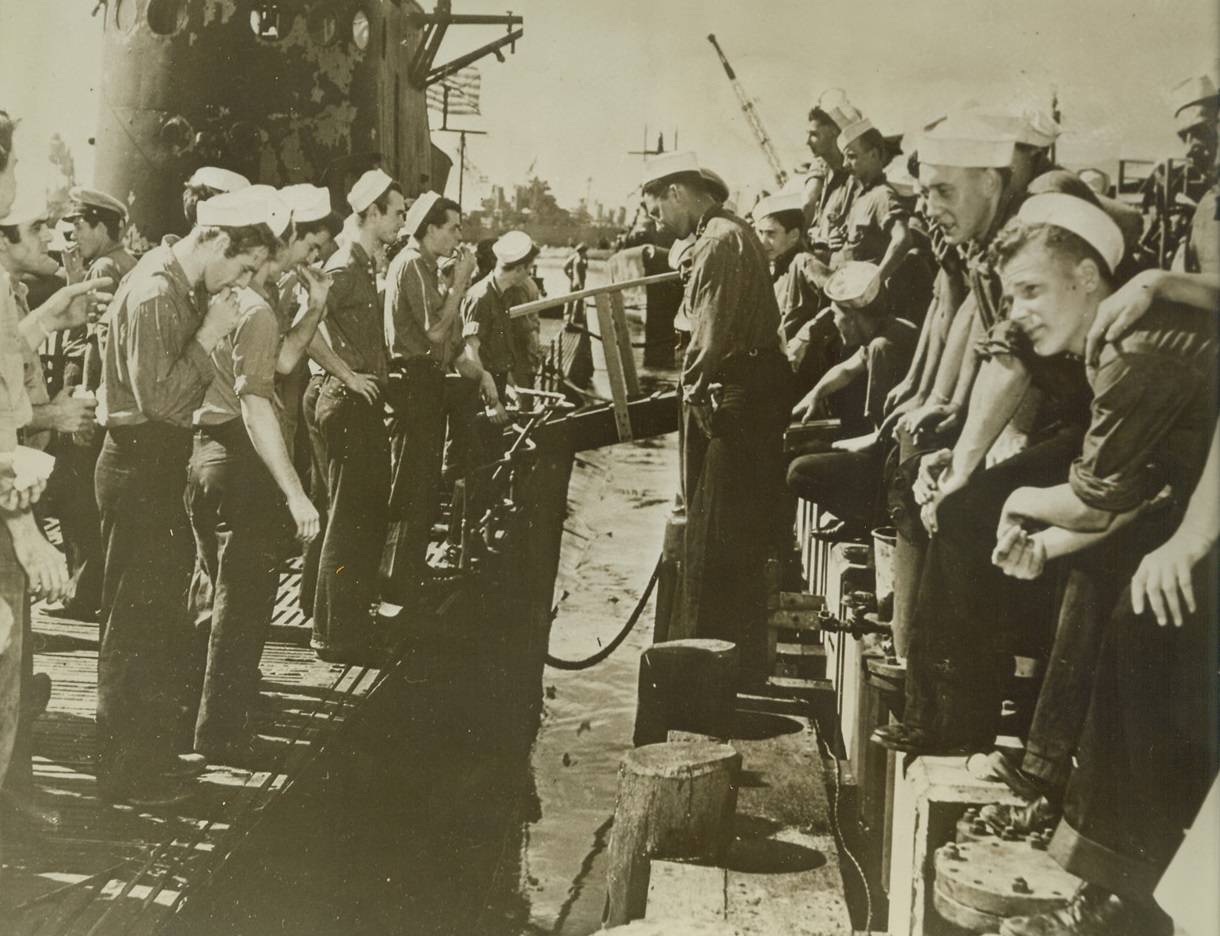 Dockside “Cracker Barrel” Session, 1/21/1943. A Pacific Base – Hungry for news, U.S. sailors line the deck of their weather-scarred submarine (left), after returning to their Pacific base, after long, arduous patrol against the enemy.  Other sailors on the dock willingly “dish the dirt” for their buddies, who have returned for rest, recreation, and, best of all, sunlight. Credit line (U.S. Navy photo from ACME);