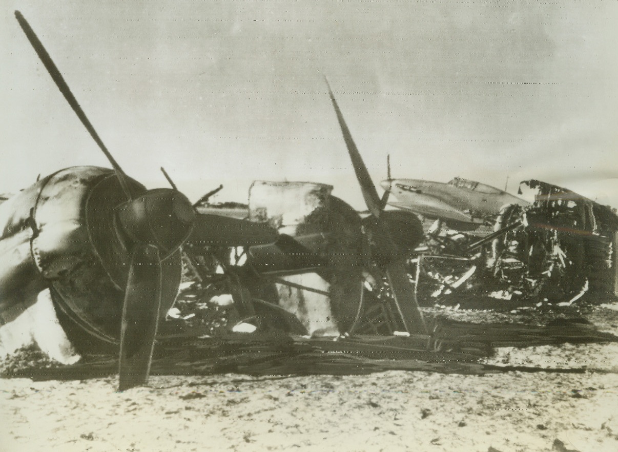 Wrecked Enemy Aircraft, 1/27/1943. Tripoli – This pile of bomb-wrecked enemy aircraft was found on Castelbenito airfield in Tripoli when allied air forces took over.  In the background, a British hurricane fighter can be seen, already operating from the captured base. Credit line (ACME radio photo);