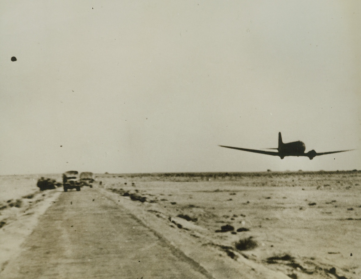 Supplies for the British, 1/23/1943. Tripolitania—A transport plane flies low over a desert road, passing a motor transport convoy as it flies with fuel supplies to RAF landing grounds in Tripolitania. Latest reports from Tripoli indicate that the city has fallen to the British 8th Army. Passed by censor.Credit: ACME;