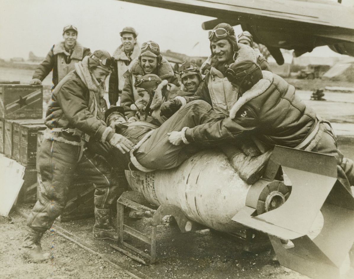 Want to Go See a Nazi?, 1/7/1943. Somewhere in England – Some of the Americans who recently shot down 19 enemy fighters in the greatest raid on Rouen threaten to fasten co-pilot Lt. Edward P. Maliszewski, of Grosse Point, Michigan, to a “block-buster.” The fighters shown here are the crew of Lt. Robert Riordan, who downed four Nazi fighters in the Rouen action. It was the second time Riordan was credited with four kills during a single battle, but he is better known for having brought his Fortress home so shot up that King George told him, “I don’t see how you did it.” This photo of the American aces at play is an exclusive ACME photo. Credit: ACME;