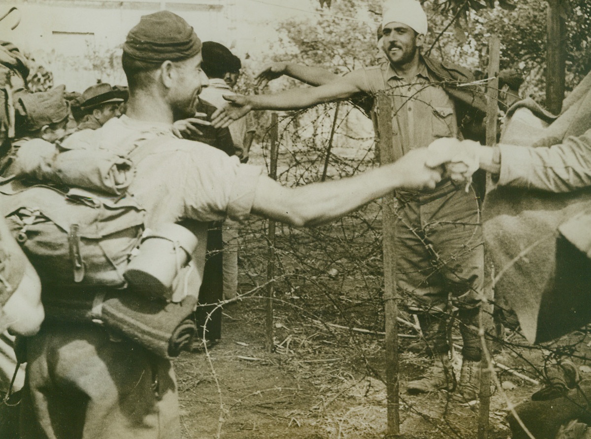 Welcome to Our Coop, 9/22/1943. Italy—As a fresh batch of Italian prisoners is brought into a temporary prisoner of war camp, their compatriots, taken in previous battles on the Italian mainland, reach across the barbed wire that forms the enclosure to welcome them. These Axis fighters were taken before their country surrendered to the Allies. Credit: ACME.;