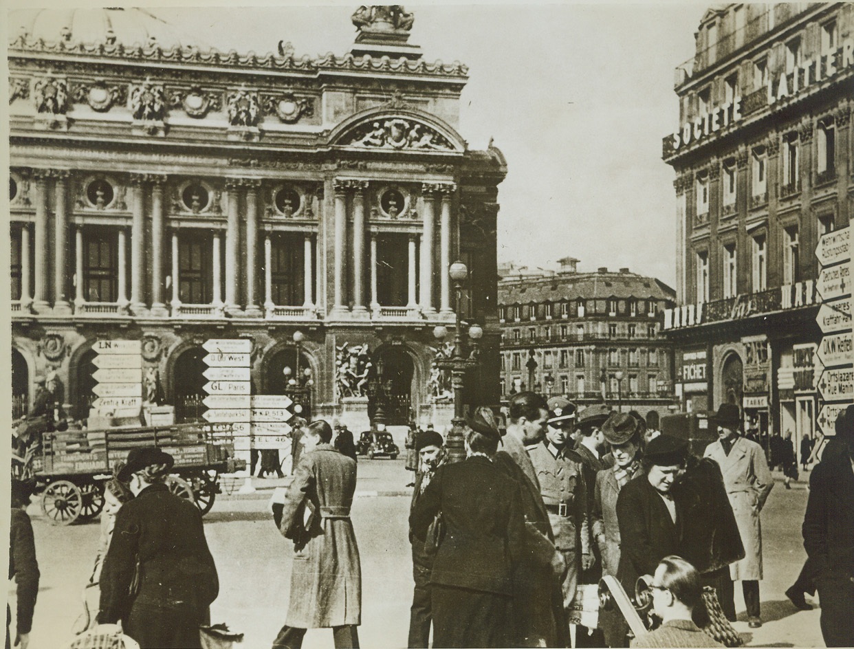 Paris Nowadays, 9/2/1943. Paris—This photo received through a neutral source shows the Place de L’Opera with the famed opera house in the background (left). Huge white sign have been placed around the square so that the “German guests” will be able to find their way around. Note Nazi officers in right foreground in midst of a group of Parisians who seem to turn away sullenly from his gaze. Credit: ACME;