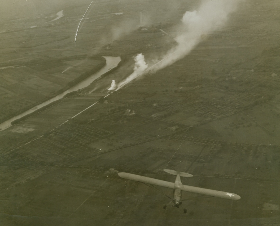 SMOKE CLOUDS THE VOLTURNO, 10/25/1943. ITALY—As artillery fire from American lines lays down a protective smoke screen to shroud coming troop movements (foreground), a different sort of smoke is visible on the Axis side of the Volturno as a result of Allied shelling. The Piper Cub is on artillery reconnaissance over German lines. Acme war photographer Charles Seawood made this aerial photo from another dub, flying between 2,000 and 3,000 feet. Credit: Acme photo by Charles Seawood, War Pool Photographer;
