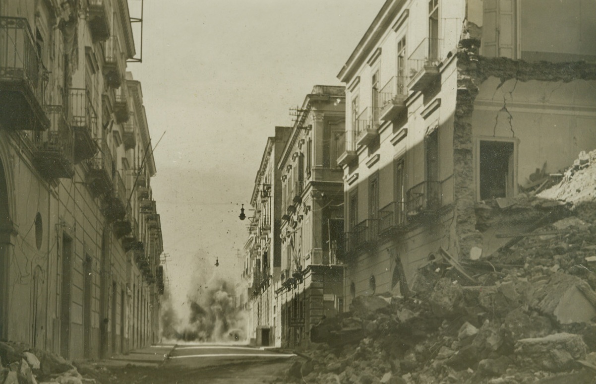 DEAD-END ROAD, 10/11/1943. ALY—A German shell bursts near the edge of Torre Annunziata at the end of a street already banked by war-wreckage. Barely visible, (left) a citizen pokes a cautious head from a doorway to see what “all the shooting is about.” Our advance on Naples was held up by such incidents as these, caused by enemy shelling, mortar bursts and snipers. Credit: Acme photo by Charles Corti, War Pool Photographer;
