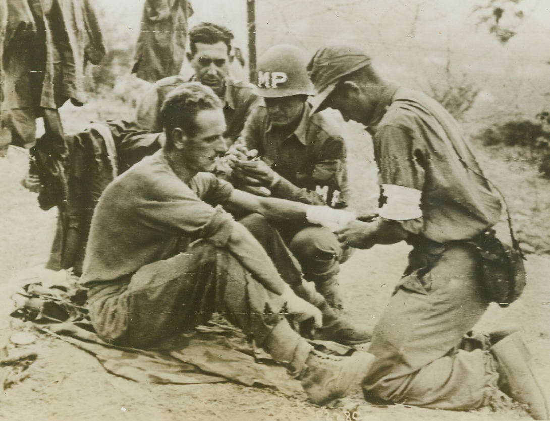First Aid for His Buddy, 10/8/1943. ITALY – Karl Kruger, a German Medical Officer from Berlin, dresses the wounds of another Nazi prisoner, E. Baecker, also of Berlin, in a stockade in Italy. Leo A. Wexler (without helmet) of Indiana, and Sgt. Anthony Cieplinzike of Chicago, Ill., both MP’s, take the names of their prisoners.Credit Line (Signal Corps Radiotelephoto- ACME);