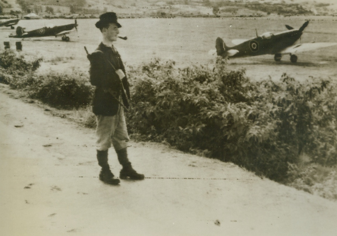 ON GUARD, 10/22/1943. CORSICA—A Corsican patriot, a gun slung over his shoulder, his pipe in his mouth, stands guard beside a group of Spitfires at an airfield in Corsica. Photo radioed to New York from Algiers today (October 22nd). Credit: R.A.F. photo via OWI from Acme;