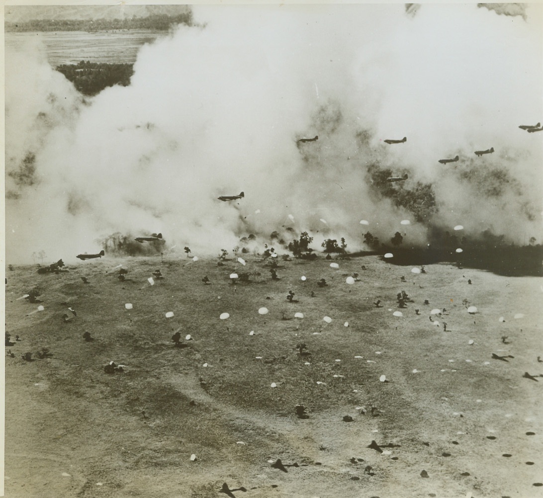 "Treetop Bail-Out" at Lae, 10/21/1943. Against a background, formed by a smokescreen laid down by Boston bombers, U.S. Army paratroopers bail out near Lae, New Guinea, from almost treetop altitude. After landing, the paratroopers captured the Jap air strip at Lae, which led to the Nipponese defeat in the area. In the photo, 11 huge American transports can be counted, many parachutes can be seen in the air, and flattened 'chutes on the ground (right, foreground) mark where paratroopers have landed. Credit: (U.S. Army Air Forces Photo from ACME);