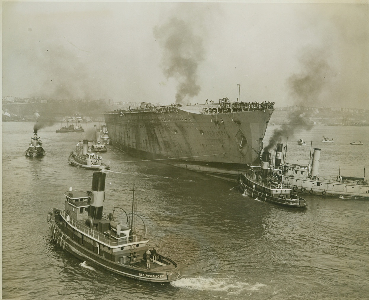 NORMANDIE HEADS FOR DRYDOCK, 11/3/1943. NEW YORK – Twenty tugs pushed and tugged the giant U.S.S. Lafayette, the former French luxury liner Normandie, down the North River when the vessel left its pier for the first time in four years, Nov. 3. Seized by the U.S. Government, almost destroyed by fire, capsized at her dock and later refloated, the great grey ship is en route to drydock at an undisclosed destination. Credit: OWI Radiophoto from ACME;