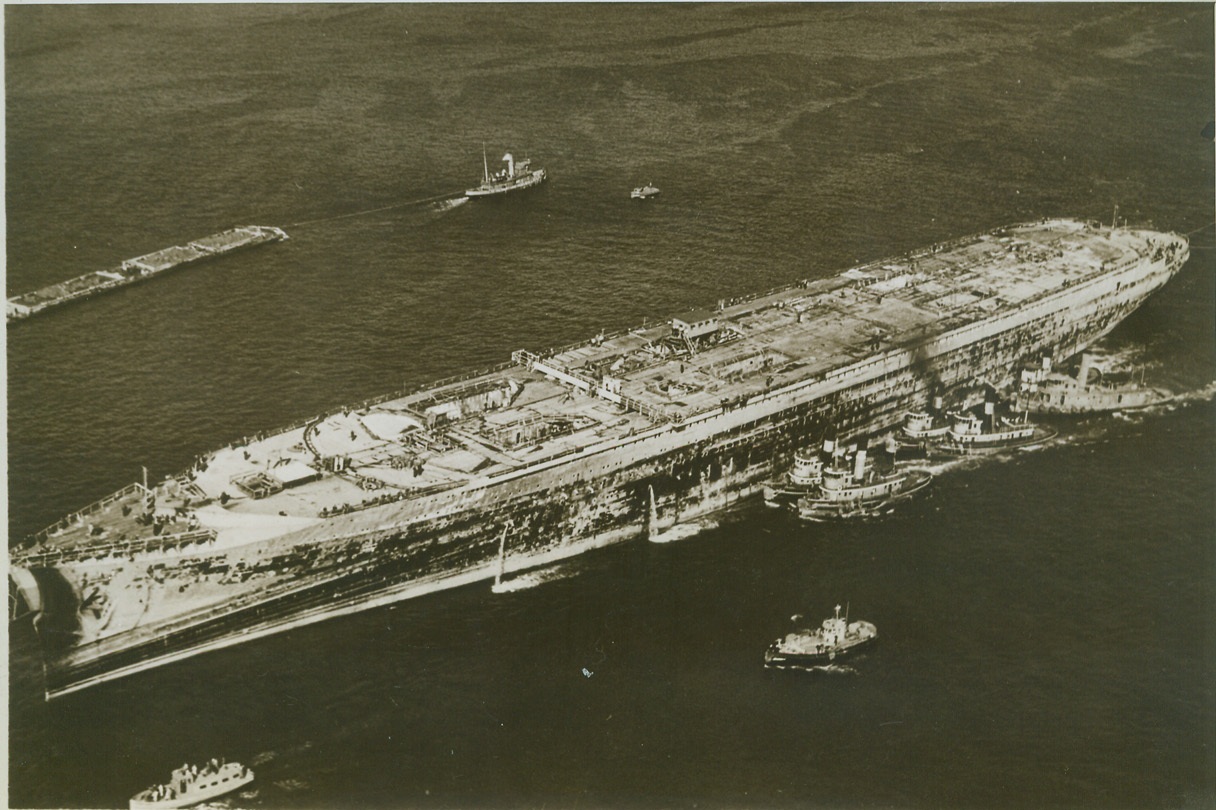 THE NORMANDIE SAILS ONCE MORE, 11/3/1943. NEW YORK, N.Y. – Her hull scarred and blackened by the fire that almost destroyed her, and her graceful superstructure gone, the U.S.S. Lafayette, formerly the French liner Normandie is towed down through New York’s Lower Bay by almost a score of puffing tugs. The vessel is on her way to drydock at an undisclosed point, where she will be refitted as a Naval Auxiliary. Credit: OWI Radiophoto from ACME;