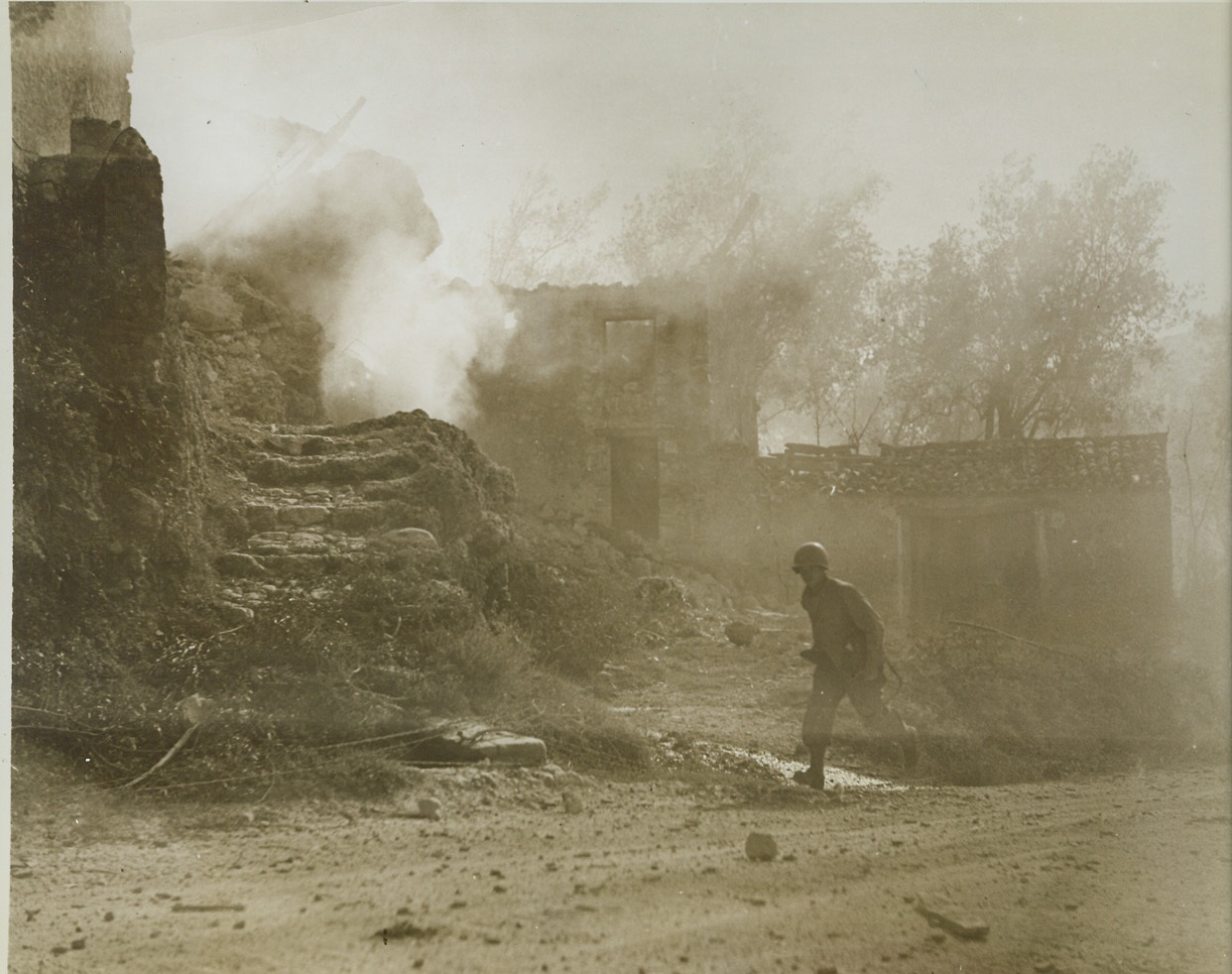 Busy Signal, 11/19/1943. Italy – A Signal Corps telephone lineman takes it on the double past a blazing house at Capriati, Italy, set afire by a direct hit from a German dive bomber. The picturesque Italian dwelling is just one more inanimate casualty of war. Credit (ACME Photo by Bert Brant, War Pool Correspondent);