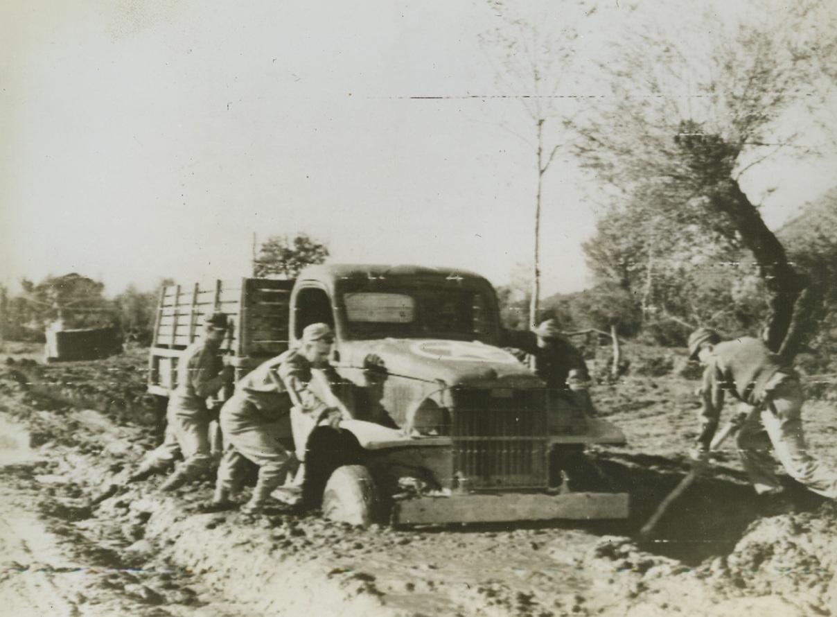 In Italy—It Rains!, 11/30/1943. ITALY—This road near the front in Italy, was one of many turned into a quagmire by recent rains, and this truck seems to be mired down for the duration. Trying to get the vehicle clear of the ooze, are (left to right): Pvt. Joseph F. White, of New Castle, Del.: Cpl. Charles R. Svoboda, Cocero, Ill.: Pfc. Ernest Shronk, (in driver’s seat), Reesburg, Wis.: Pvt. Murray Marks, Bronx, N.Y.; and Cpl. Cecil Cox, (with pole), of Bloomington, Ind. Credit: U.S. SIGNAL CORPS RADIOTELEPHOTO from ACME.;
