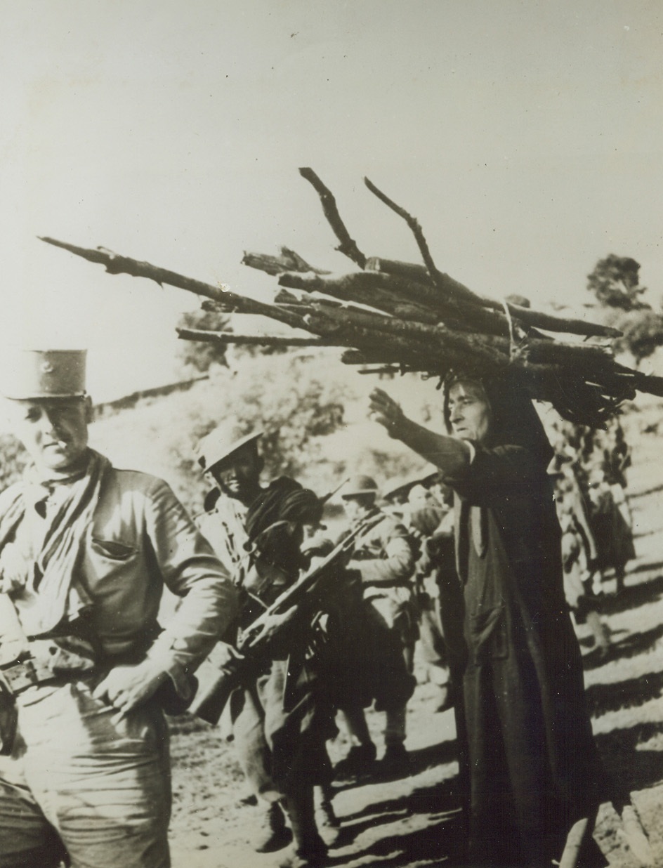 POINTING THE WAY, 11/12/1943. CORSICA—Pausing as she collects firewood and balancing a huge pile of the fuel on her head, an aged Corsican woman gives directions to French soldiers. Natives cooperated wholeheartedly with the warriors, who soon drove the Nazis out.Credit: French official photo thru OWI from Acme;