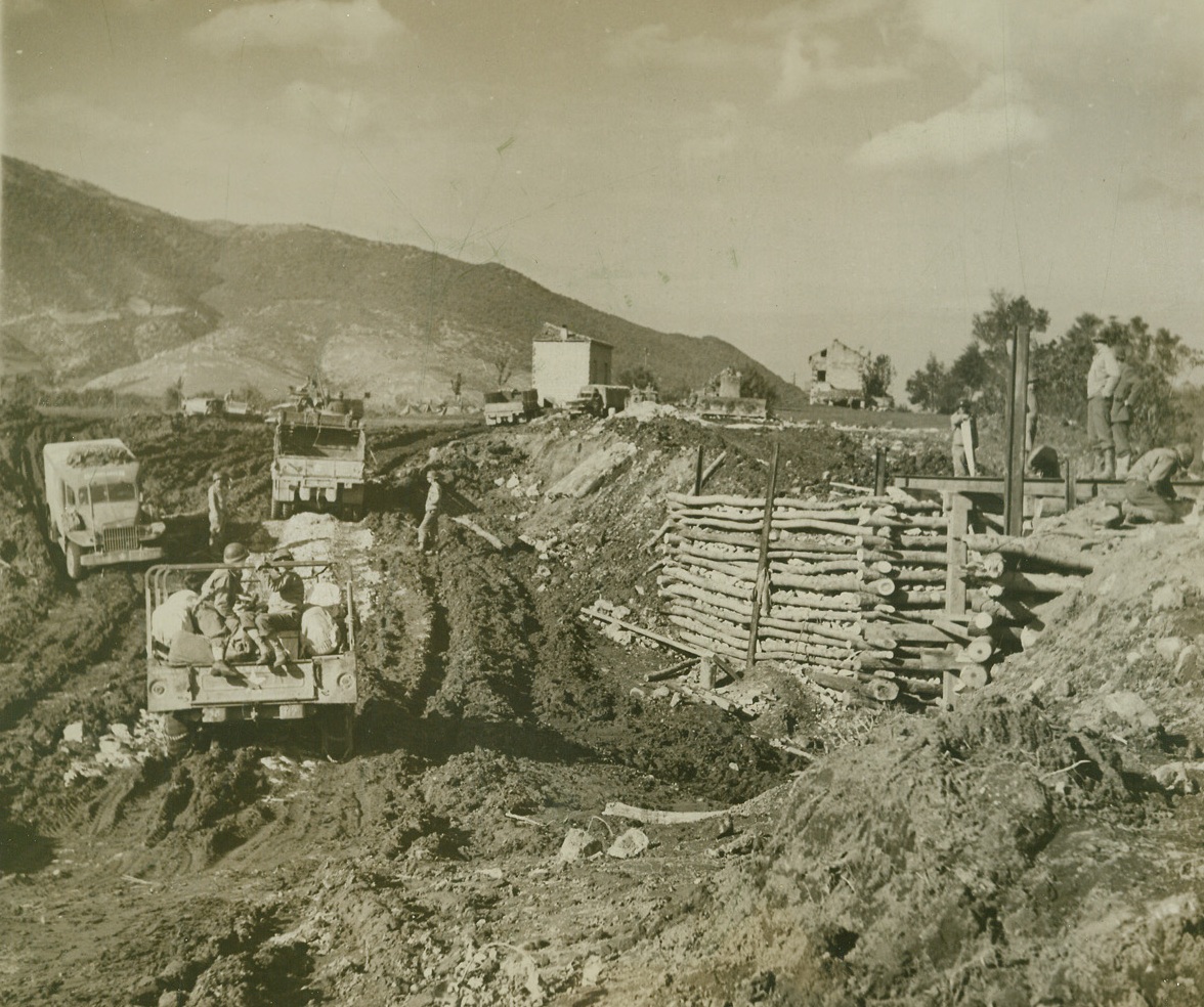 Battlefront Boulevard, 11/23/1943. WITH THE FIFTH ARMY IN ITALY -- Jeeps and trucks of the U.S. Army, plow through hub-deep mud crossing this dry stream bed on a temporary road, along a section of the Italian Front. In center, background, engineers work to complete a bridge to replace one blown up by the Germans as they retreated to Cassino. Credit Line (ACME Photo by Bert Brandt for the War Picture Pool);
