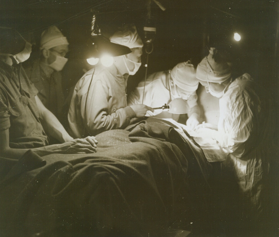 Men in White - At the Front, 11/28/1943. SOMEWHERE IN ITALY -- Makeshift lighting casts an eerie glow about this "operating room" as white-clad Army surgeons perform a difficult operation in a field hospital behind the front lines. The strain of their work increased by inadequate facilities, these heroic doctors render immediate surgical treatment to men picked off the field, who are too far gone to withstand a trip to an evacuation hospital before the operation. Credit Line - WP - (Photo by Bert Brandt, ACME Correspondent for War Picture Pool);