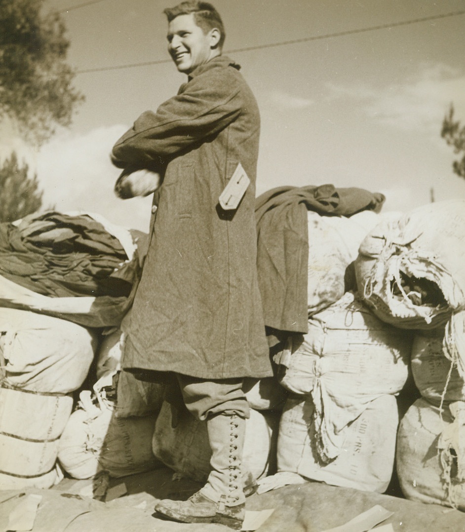 G.I. FASHION PLATE, 11/23/1943. WITH THE 5TH ARMY IN ITALY—Pfc. Thomas Patrick, of Hilton, Okla., tries on a warm G.I. winter overcoat, which will become an indispensable part of his equipment during the cold Italian winter ahead. Behind him (in photo) are bales of coats, warm gloves, winter underwear, etc., being passed out to Americans on the 5th Army front in Italy. Credit Line (ACME Photo by Bert Brandt for the War Picture Pool);