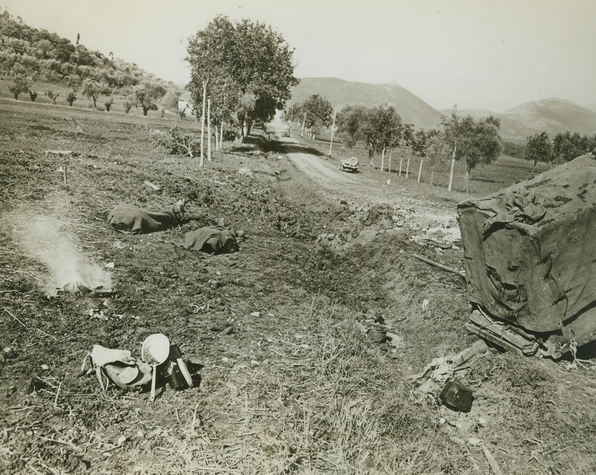 Death Marks the Spot, 11/16/1943. Italy – The bodies of two American soldiers (left background), who had been riding along this road in Italy in a half-track, lie beside the road after a German air attack had blasted the motor vehicle to wreckage and killed them. The bomb crater can be seen at right just behind the battered trailer. Bits of wreckage, mess gear, and helmets litter the ground and a burning bit of wreckage, (left) marks the scene of death. Credit: U.S. Army official photo from ACME;