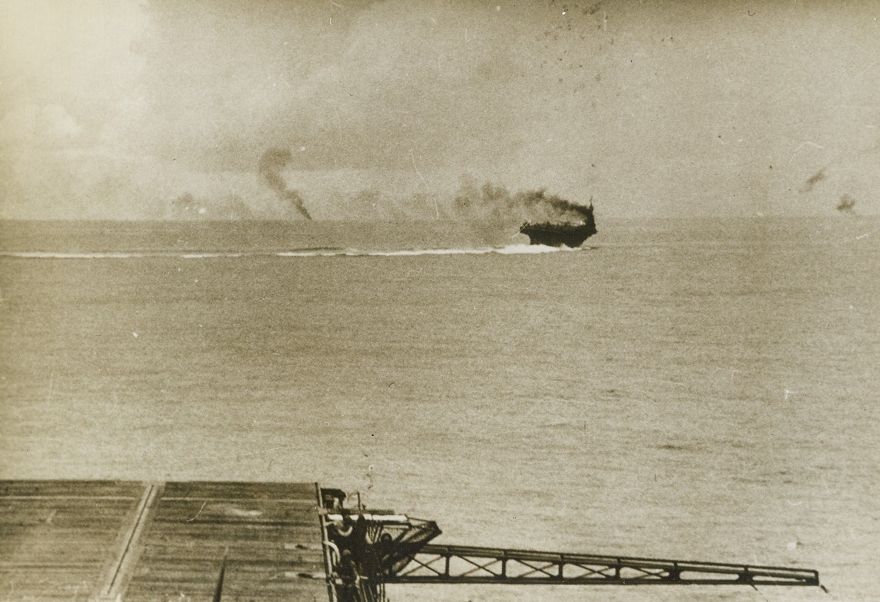 Yank Flat-Top Outwits Japs, 11/26/1943. South Pacific – With the deck of one U.S. Aircraft Carrier in the foreground, another of our Flat-Tops maneuvers wildly to escape Jap bombs while (left background) a Jap plane falls into the sea in flames, during the spectacular U.S. attack on Rabaul when our carrier-based planes shot down 70 Nip aircraft and sank and damaged two Jap Cruisers and 13 Destroyers. With the sky raining bombs and blazing Japanese planes, none of our ships were hit. Credit: ACME;