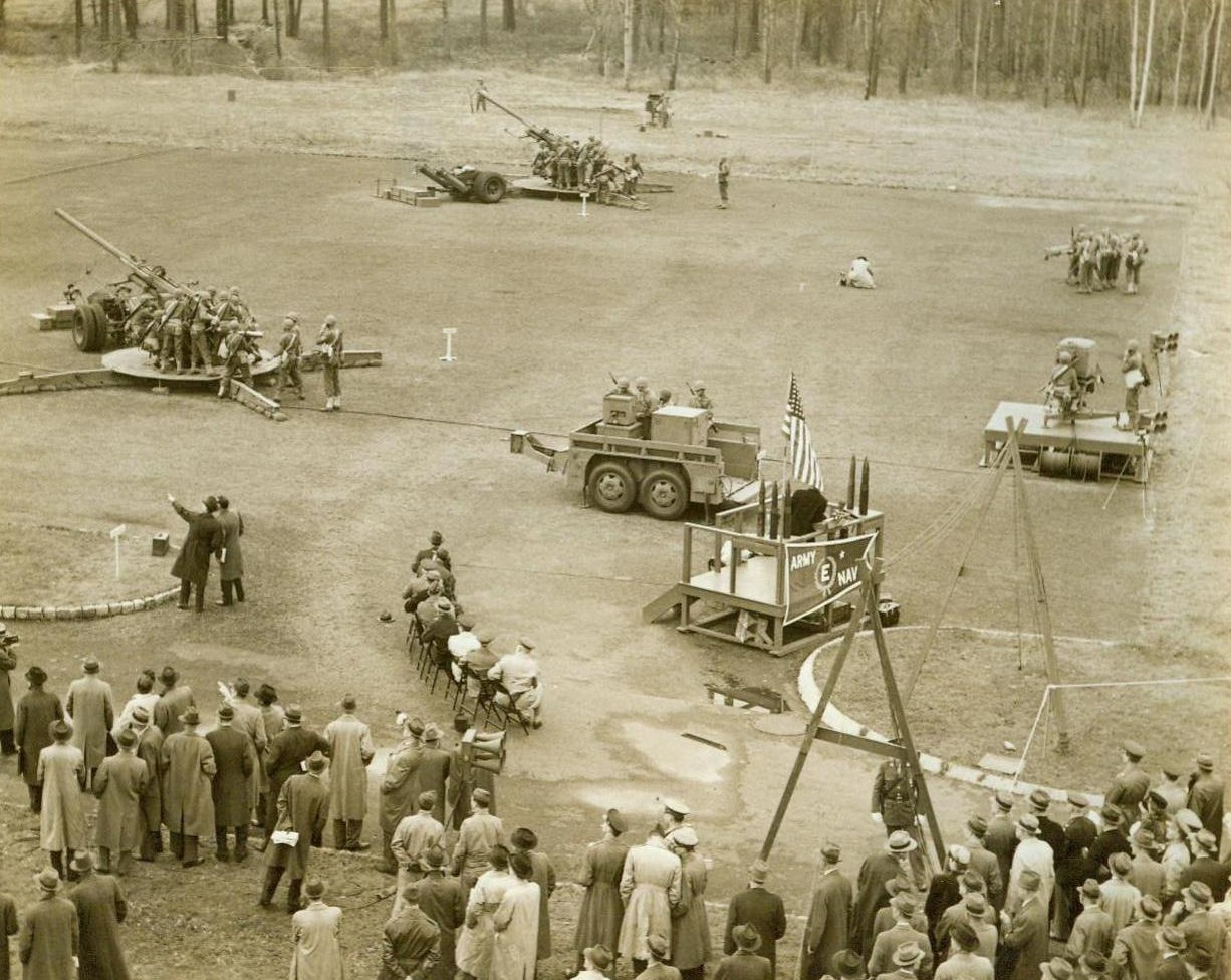 Electrical Impulse Guides Ack-Ack Shells, 11/10/1943. Murray Hill, N.J. -- Here is a general view of the Army demonstration of the new M9 gun pointer, shown at the Bell Telephone Laboratory in Murray Hill yesterday. At left are two 90MM anti-aircraft guns; in center, to the left of flag, is the electrical computer which directs the guns; in right center is tracker unit and in right background is the height finder. 11/10/43 (ACME); in right center is tracker unit and in right background is the height finder. 11/10/43 (ACME);