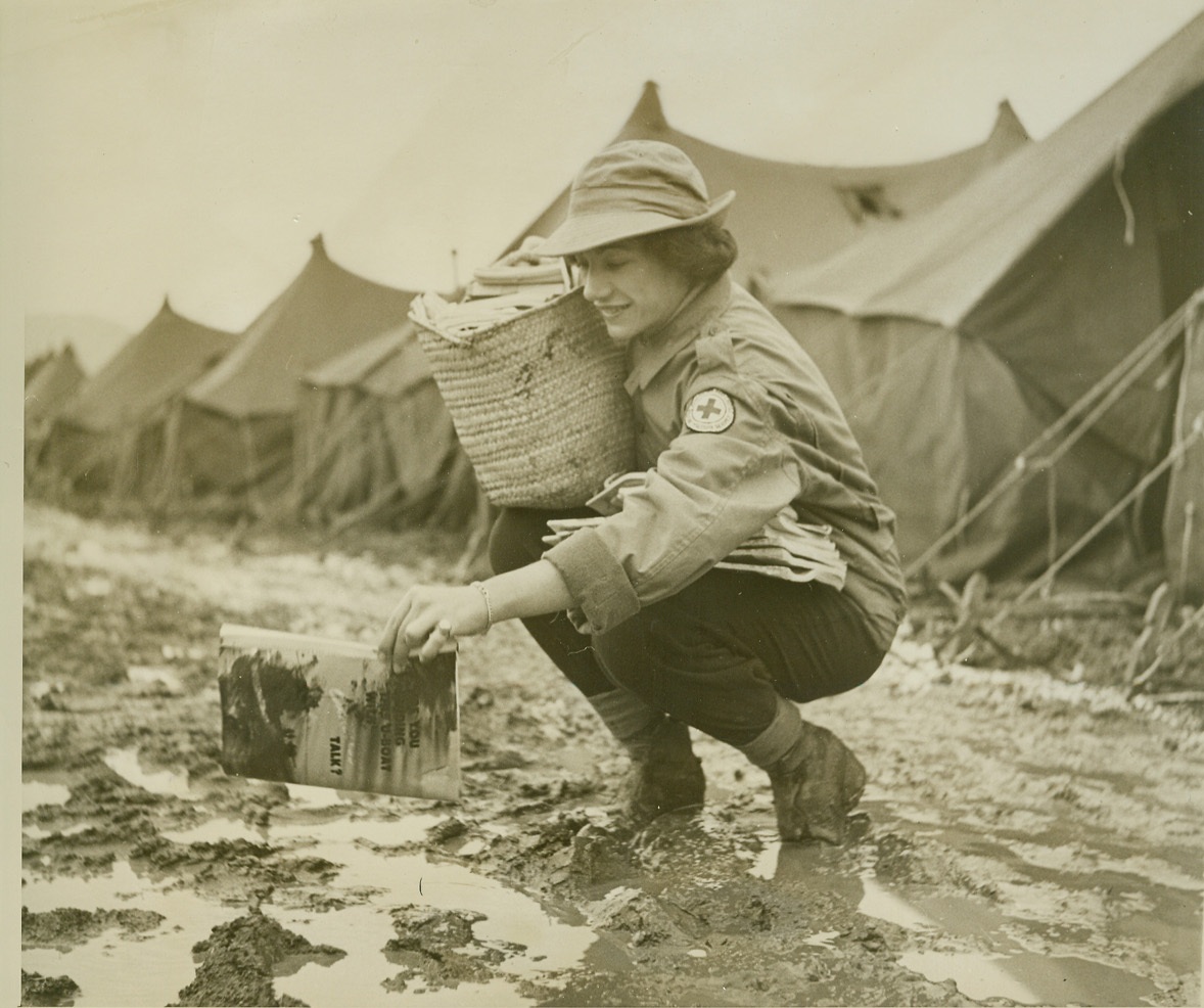 Magazine Bites the Mud, 12/3/1943. Somewhere in Italy – “Butterfingers” don’t go on the Italian front these days because anything dropped in the mud that covers the countryside is ruined. Red Cross worker Nancy Gres of Narberth, Pa., stoops to pick up a mud-covered magazine dropped as she was making the rounds of an evacuation hospital with books, cigarettes and magazines. Credit: (Photo by Bert Brandt, ACME Correspondent for War Picture Pool);
