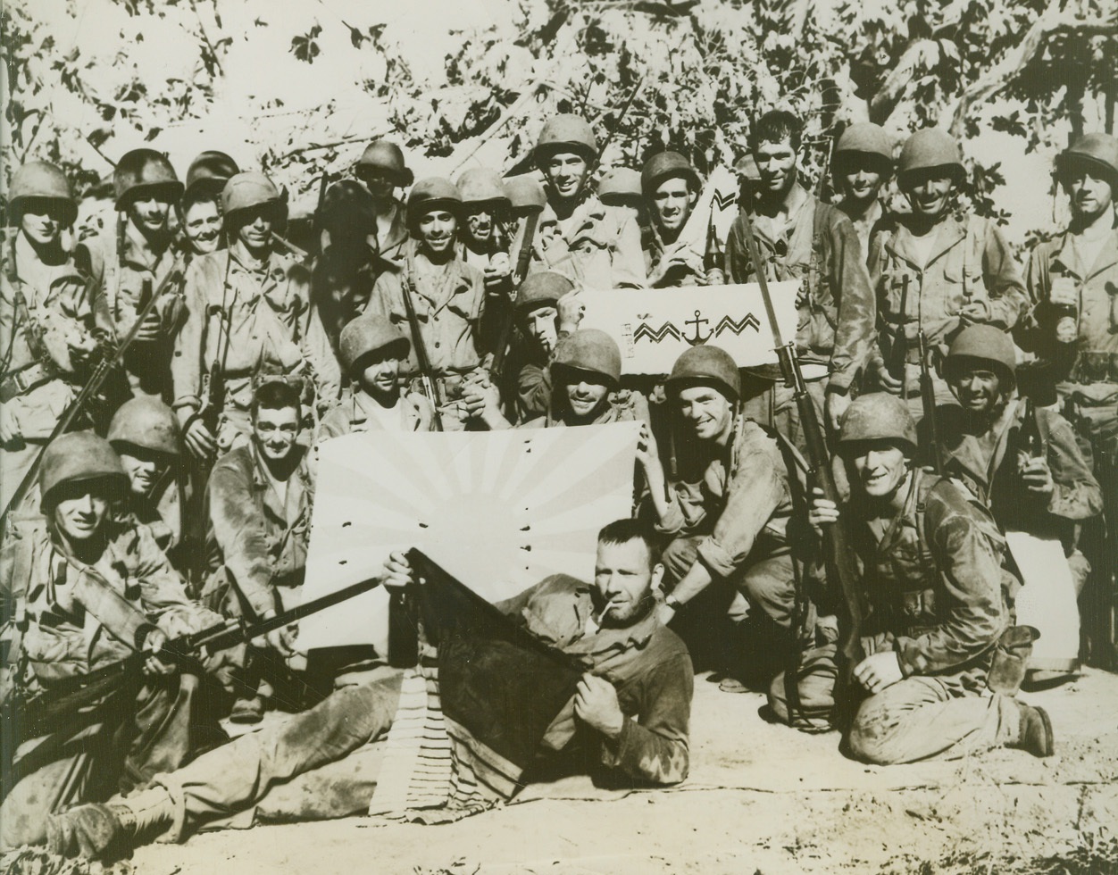 DISPLAY OF CAPTURED SOUVENIRS, 12/9/1943. MAKIN ISLAND—Back from the battle front with their captured souvenirs are the men of the 165th Infantry, the old “Fighting 69th”. Pictured are a Japanese flag and Japanese marine insignia taken at [illegible] grove, Makin Island.Credit: Official U.S. Army Signal Corps photo from Acme;