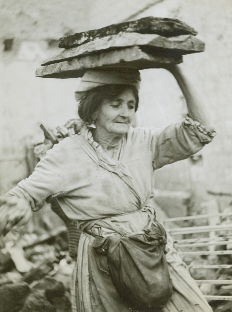 Precious Burden, 12/22/1943. Mignano, Italy—Walking unsteadily through the ruined streets of Mignano, this old Italian woman carries a precious burden atop her head. The three huge slabs of wood, found in the bomb debris that clutters the town, will be used to provide fuel for her cookstove. Credit: Photo by Sherman Montrose, ACME photographer for War Picture Pool.;