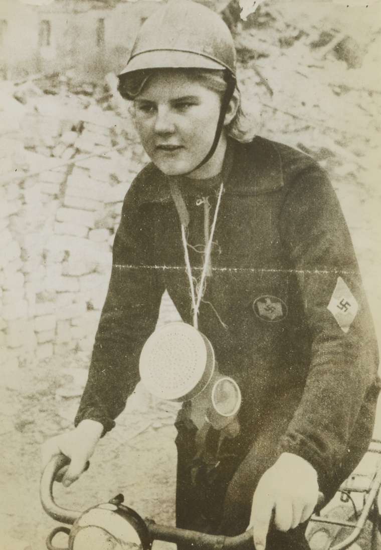 Berlin Air Raid Warden, 12/13/1943. BERLIN—A young girl, obviously still in her teens who is probably on of Berlin’s hastily enrolled air raid wardens, rides through the city’s blitzed streets on a bicycle the morning after an all-out Allied raid. She is equipped with a helment, gas mask and overalls. Note Swastika on her arm and pile of debris in the background. Photo received through a neutral source. Credit: ACME.;