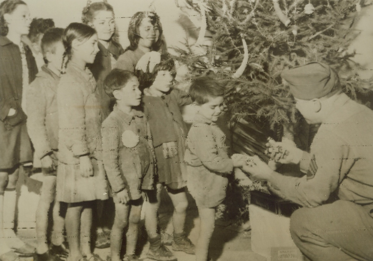 MERRY CHRISTMAS FROM THE YANKS, 12/26/1943.ITALY—The Christmas spirit is not swallowed up in war in Italy, either, where Sgt. Patsy Di Custanzo, of New York City gives candy to native youngsters in Caserta. Although goodies are scarce in their country, the Italian children patiently await their turn for the Yankee gifts.Credit: OWI Radiophoto from Acme;
