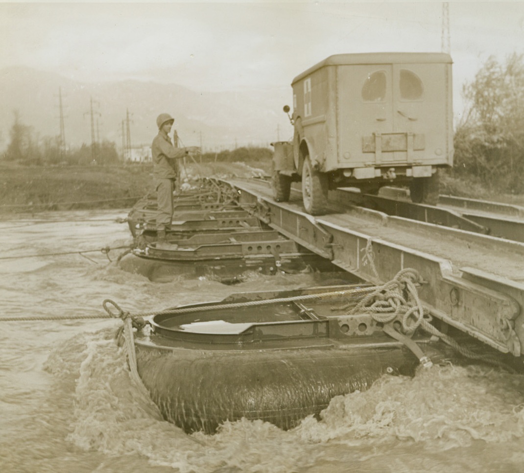 The Roaring Volturno, 12/8/1943. ITALY – High, rushing waters of the Volturno River, swollen by recent heavy rains, churn around pontoons of bridge set up by U.S. Army Engineers. Flood waters had wrecked a least one pontoon bridge. Pvt. Edwin Reidler, Reading, Pa., is giving the high sign to traffic. Note heavy ropes which keep pontoons in place. Credit Line – WP – (ACME);