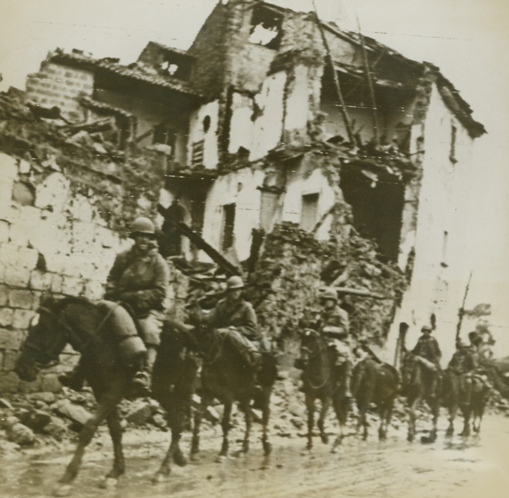 WHERE THE RAIN REIGNS, 12/22/1943. ITALY—In Italy, where the biggest enemy is bad weather, a soggy unit of U.S. cavalry rides past a shell-torn building on the Italian front. Allies have been borrowing Italian horses for reconnaissance missions over narrow, slippery lanes impassable to mechanized vehicles. Credit: Acme photo by Bert Brandt, for the War Pool via Army radiotelephoto;