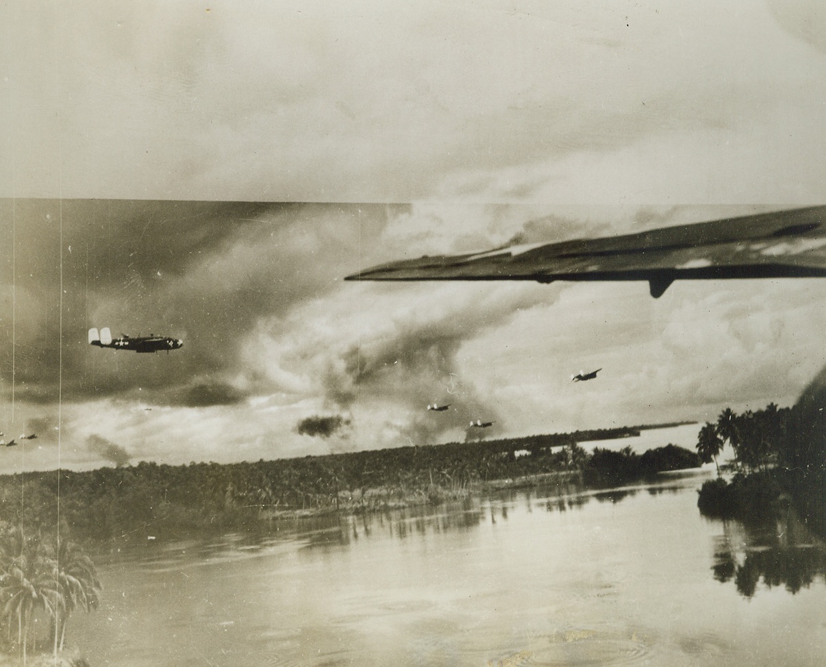 Yanks Blast Madang, 12/10/1943. New Guinea – Low-flying B-25s run over the target and fairly skim the water of the bay at Madang, New Guinea, where columns of black smoke in the background mark destruction caused by American B-24s on Alexishafen. The black puff of smoke in the center of the photo is from enemy ack-ack. Credit: ACME;