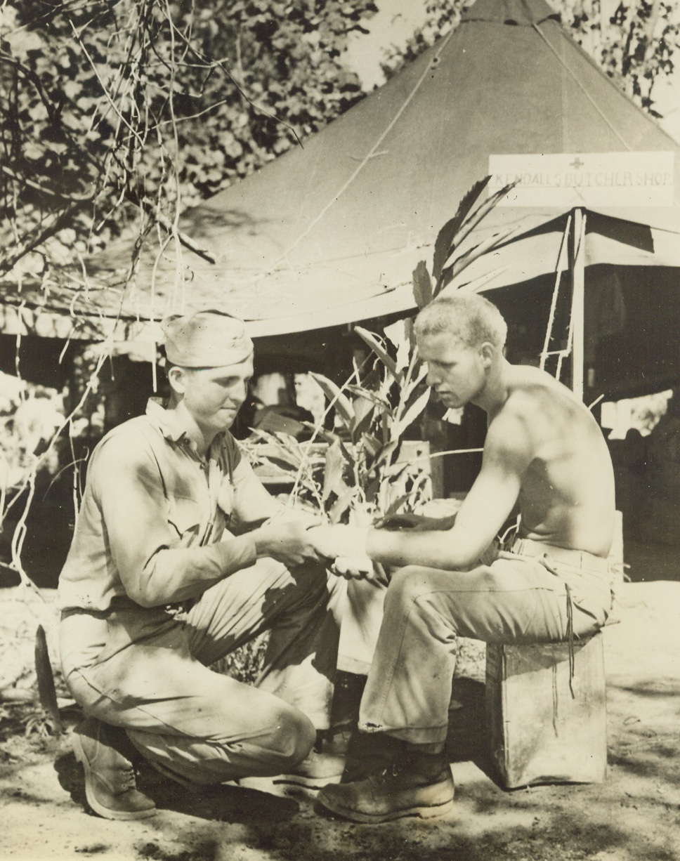 Hero Carries On, 12/30/1943. Vella Lavella, S. I. – Pharmacist’s Mate 2/c Rex H. Gregor, 21, of Rochester, Minn. (left), recently performed one of the most heroic acts of the war when he climbed aboard a blazing Landing Barge on which ignited ammunition was exploding, helped remove the wounded and then performed a leg amputation on one of the casualties – although Gregor had never seen such an operation. The action occurred during a Japanese air raid on Marine forces on Vella Lavella, in the Central Solomons. Here, Gregor bandages the wrist of Cpl. Kenneth A. McIntyre, USMC, of Minneapolis, Minn., who also helped rescue wounded from the blazing barge. Note sign over tent, (top right), reading, “Kendall’s Butcher Shop.” Credit: Official Marine Corps photo from ACME;