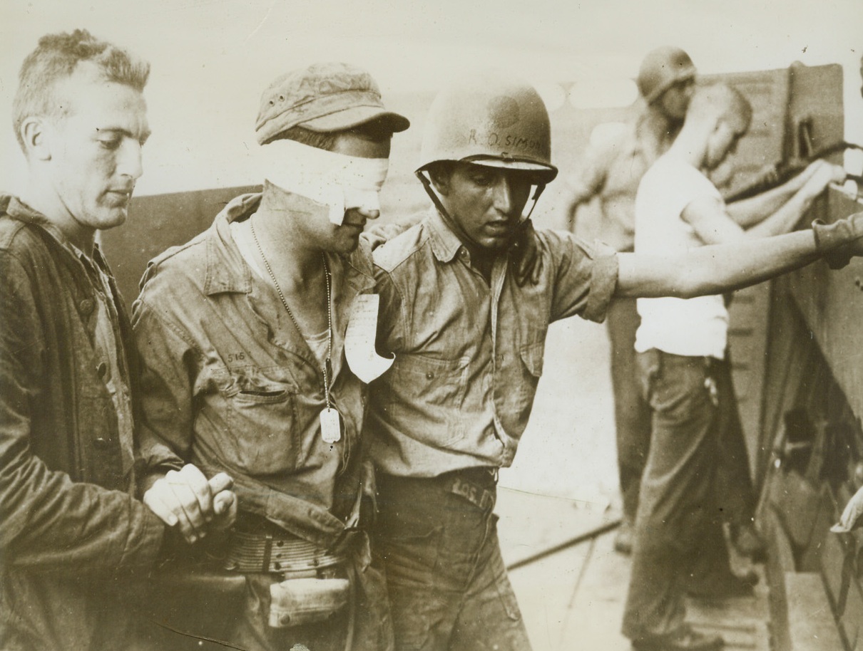 A Yank Leaves Makin, 12/5/1943. MAKIN—Flying enemy shrapnel found the eyes of this U.S. Army private, who wears bandages as he leaves Makin Island. Two medical corpsmen assist the Yank, who helped our forces gain possession of the central Pacific atoll, to board a transport. Credit: ACME;
