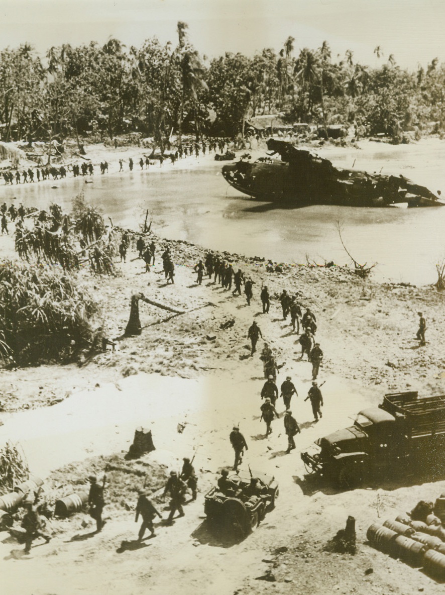 Out to Get What’s Left, 12/18/1943. Makin Island – Out to get what’s left of the Jap force pushed back to one end of Makin Atoll, a column of American fighters tramps around a lagoon in which a Japanese seaplane is partially submerged after strafing by Yank planes.  In the right foreground are stacks of Jap fuel barrels. Credit (U.S. Coast Guard photo from ACME);