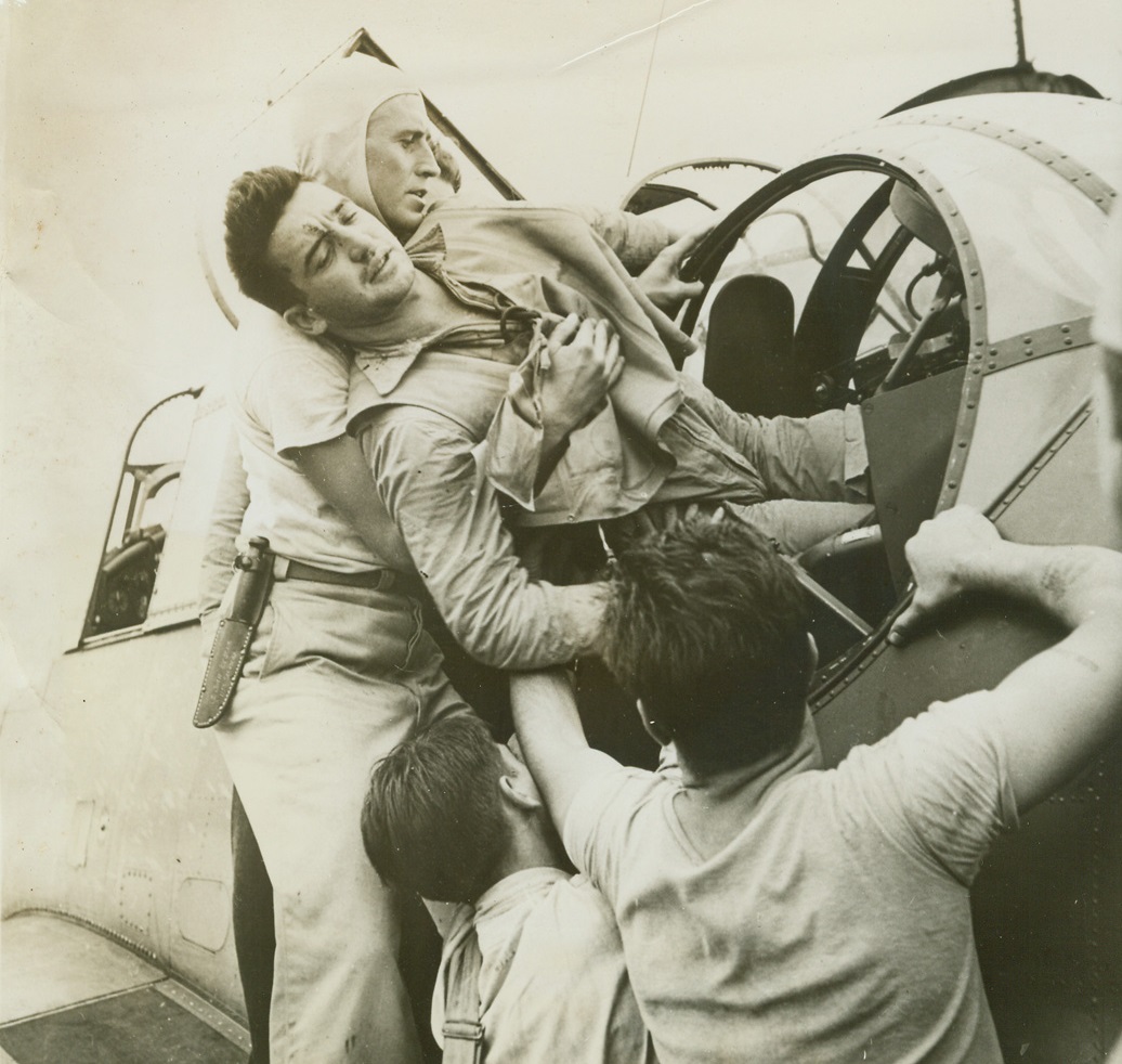 Raiders’ Return, 12/16/1943. At Sea – Returning to the flight deck of the USS Saratoga after the terrific, November 5th raid on Rabaul, wounded rear gunner AOM Kenneth Bratton, of Mississippi, winces as he is lifted from the turret of the Avenger in which he fought. Applying a tourniquet to his shattered knee, Bratton managed to retain consciousness until he was carried from his post by Lt. Julie Bescoes, USNR, formerly a University of  California coach and All-American grid star. 12/16/43 Credit Line (Official U.S. Navy Photo From ACME);