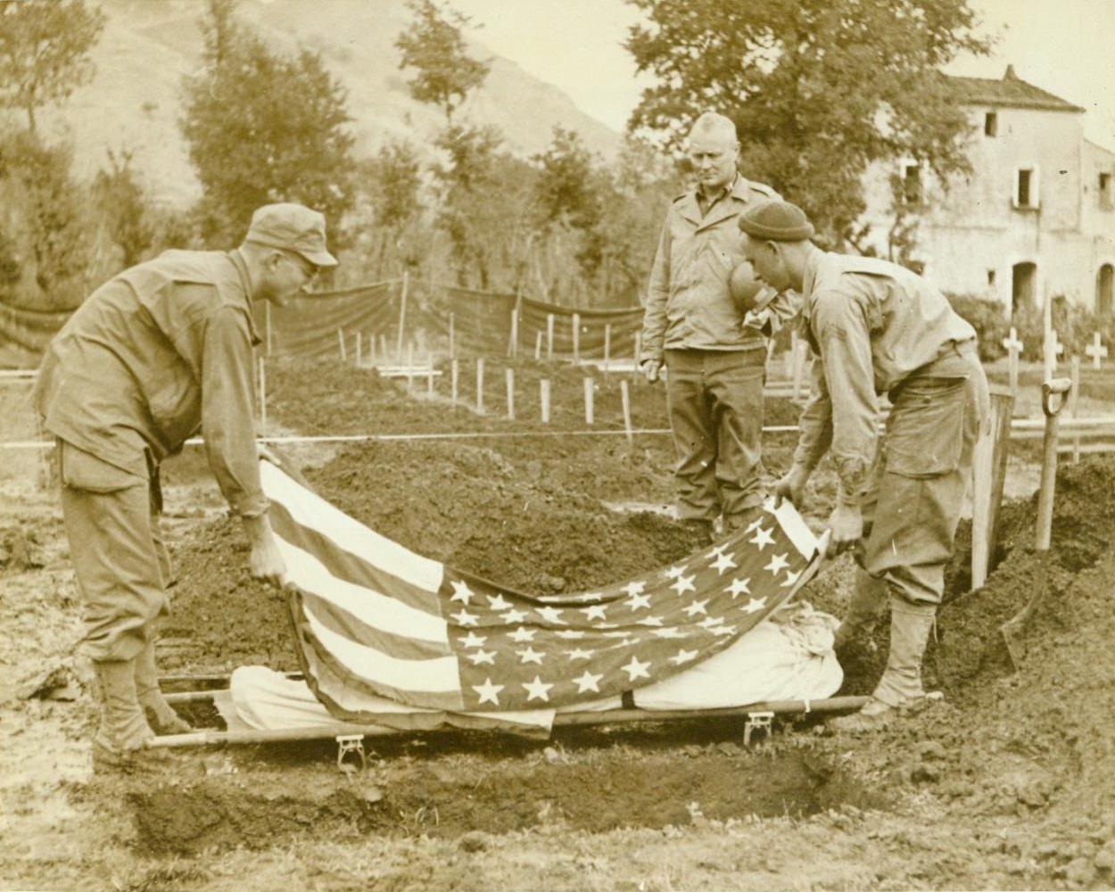 Last Rites in Italy, 12/19/1943. Italy—Capt. Christ A. Lehne, Lutheran minister from Fredericksburg, Texas, stands with bared head as two Fifth Army Fighters remove the stars and stripes from the body of a dead comrade. In the background, temporary markers and white crosses mark the resting places of other Americans who gave their lives in Italy. 12/19/43 (Acme);