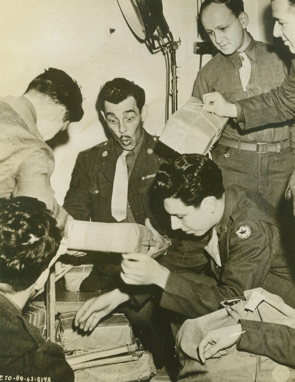 It’s Christmas for Yanks in Britain, 12/13/1943. England – The arrival in England of Christmas packages for Americans soldiers brings cheer and anticipation to this group of Yanks. Sgt. Kenneth L. Thiem (center, facing camera), of Los Angeles, California, whistles with delight as he receives his package, while Pvt. Leslie Rachline (kneeling, right) of North Bergen, New Jersey, eagerly searches for his gift. At top right (left to right) are Pvt. Mitchell Kucharsky, Brooklyn, New York, and Pvt. Walter W. Newman, Fort Wayne, Indiana. Credit: U.S. Signal Corps Photo from ACME;