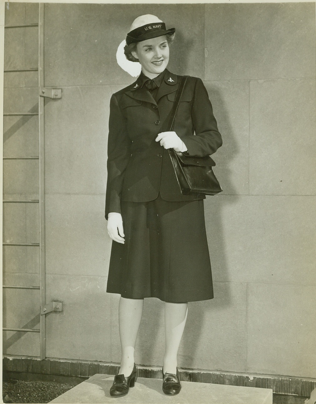 Uniform of WAVES’ Enlisted Personnel, 10/1/1942. NEW YORK – The enlisted personnel of the WAVES will wear a uniform exactly like that of their commanding officers’ except that black buttons take the place of gold, and rating marks on the upper sleeve supplant gold braid. The skirt is exactly the same, and the hat has a six section crown with wide stitched brim and a white crown for summer wear, replaced by a blue crown for winter. Both hats have a black band with “U.S. Navy” in gold. The Mainbocher designed uniform is modeled by Miss Ellen Allardice. Credit: (ACME);