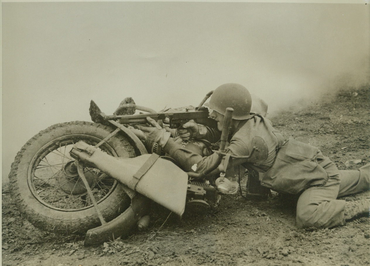 Two Uses for Motorcycle, 10/13/1942. ENGLAND -- Corp. Francis Rivers, of Cambridge, Mass., one of scouts of "attacking" forces during maneuvers of American troops in England, uses his motorcycle as a shield while firing at the "enemy." During these maneuvers, the use of chemical warfare was stressed for the first time.  Credit: (ACME);
