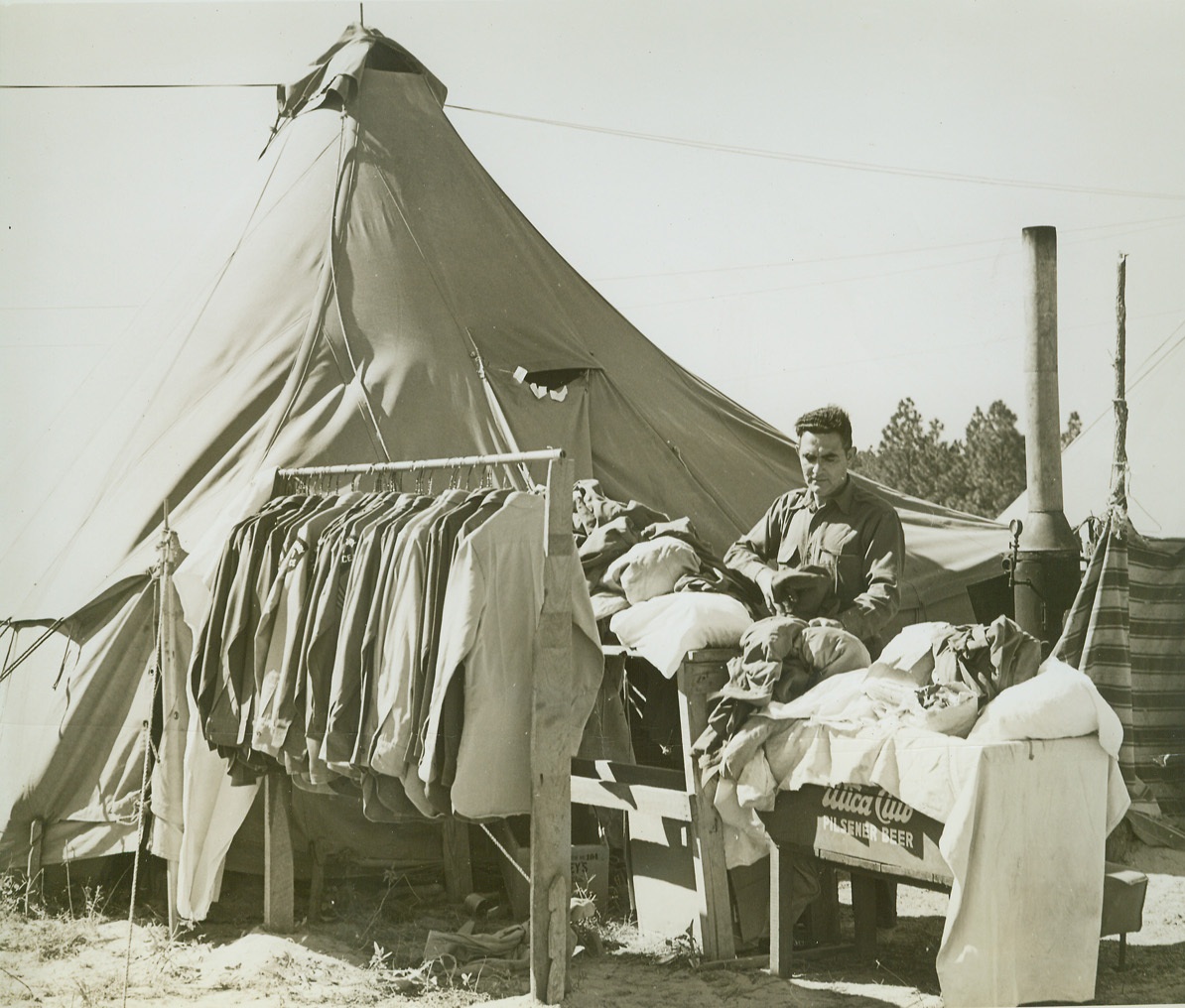 FIELD TAILOR SHOP, 10/26/1942. MANESS, N.C. – Irving Levinson of New York, who has just put out a rack of freshly pressed uniforms for special troops of the 26th Division, goes to work assorting a new batch of laundry and uniforms. A civilian tailor, Levinson’s shop is now a tent in the camp area. In background right is the kerosene fired steam boiler for pressing. Credit: OWI Radiophoto from ACME;
