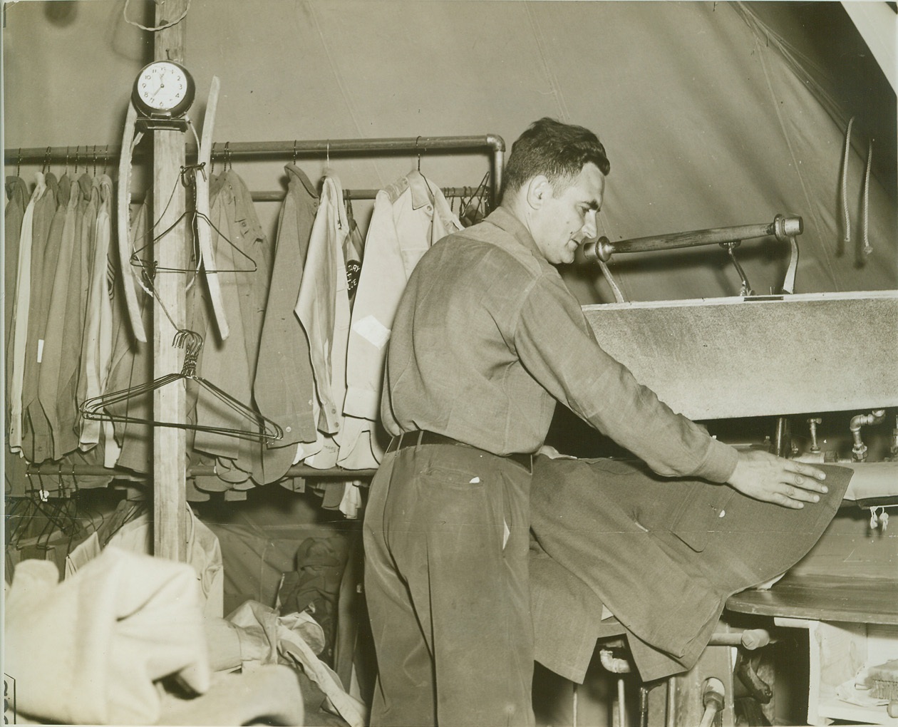 FIELD TAILOR SHOP, 10/26/1942. MANESS, N.C. – Irving Levinson, a tailor by trade, keeps in trim caring for special troops of the 26th Division. His new shop is a tent in the camp area. Here, the New Yorker does some pressing. A kerosene fired steam boiler operates the presser. Credit: OWI Radiophoto from ACME;