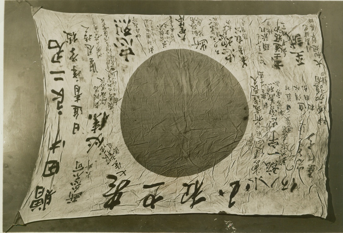 Jap Battle Flag Taken by U.S. Correspondent, 10/27/1942. NEW YORK CITY – Prize trophy from Guadalcanal of United Press Correspondent Robert C. Miller is this slogan-covered Jap battle flag which the newsman took from a Jap officer (presumably a colonel) slain in the Battle of Tenaru River on August 21st. Miller, who landed with the first wave of U.S. Marines at Guadalcanal and remained there for the first 6 weeks of occupation, got the permission of the Marines’ intelligence department to send the flag to New York to be used to raise funds for the war effort. Large inscription nearest staff side of the flag reads: “Presented to Tanak Rejoji, Our Buddy.” Next large line from the staff reads: “ Japanese Progressive School for Young Men.” Other legends read: “Heads Up,” “Be Courageous,” “Fight to the Finish,” “Bravery is Patriotism,” “With Great Courage,” “Cherry Tree for Flower, Man for Warrior,” and “Bushido.” Credit: (ACME);
