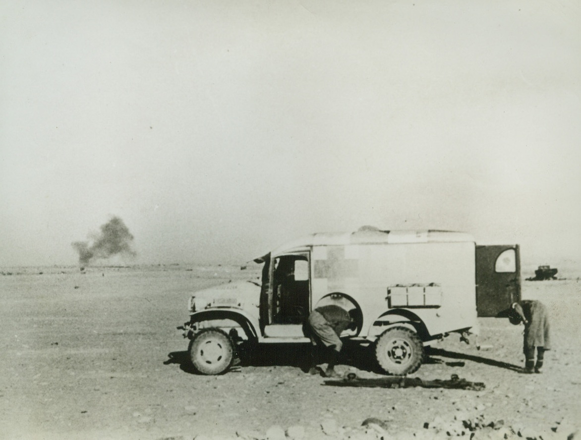 Ducking Shrapnel, 10/25/1942. North Africa—Edward J. Sullivan, ambulance driver from Kansas City, crouches beside his vehicle as shells burst nearby. A New Zealander takes refuge behind the open door as they wait for a lull in the fire to go out and pick up possible wounded. Both are with the American Field Service, which provides ambulances for Allied forces in Africa. Credit: ACME.;