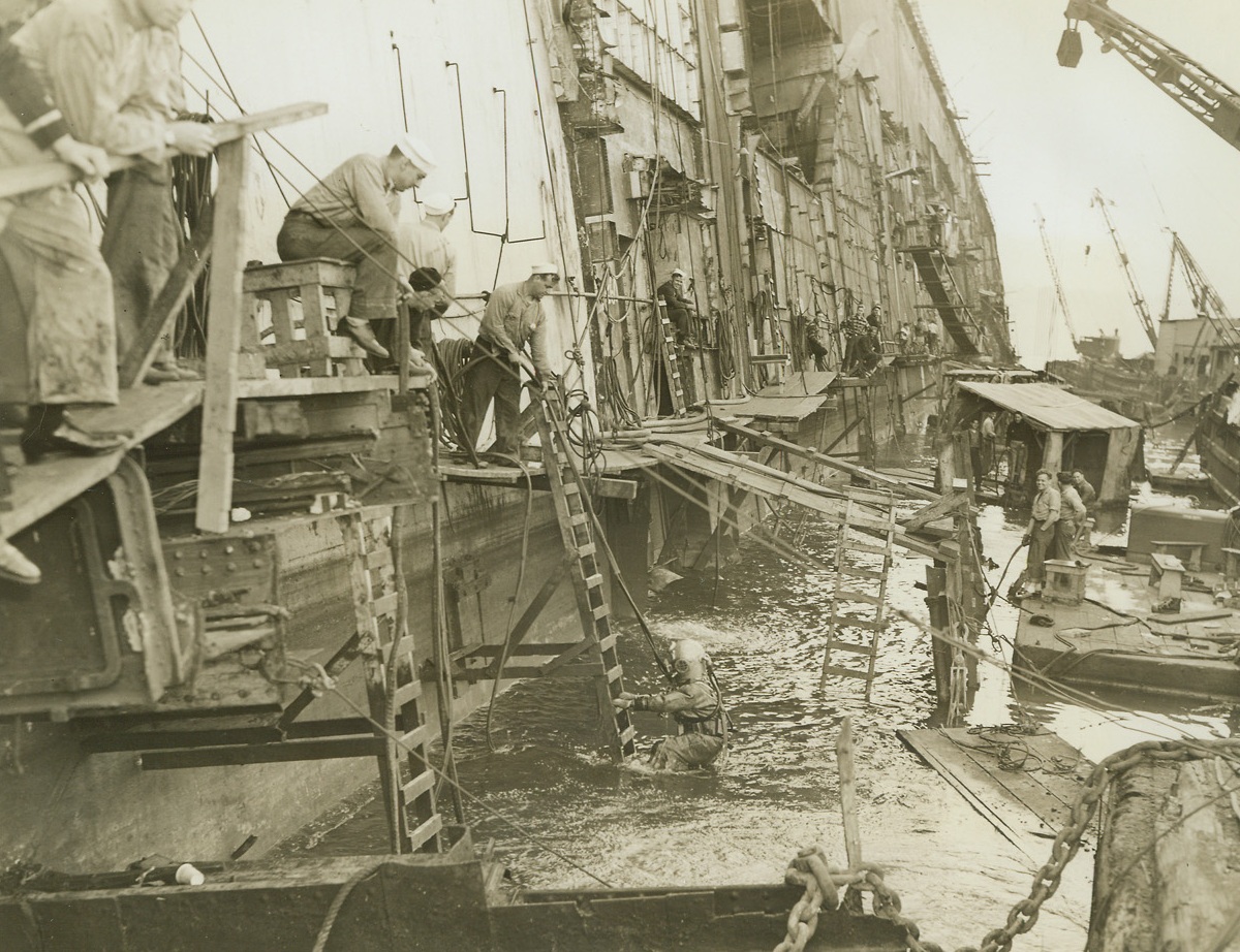 No Title. 10/9/1942. Many men are at work along the catwalk of the S.S. Lafayette, as a diver comes to the surface and is aided up a ladder to the walk. Cranes, rafts, and a maze of temporary structures surround the grey ship’s hulk.Credit: ACME.;