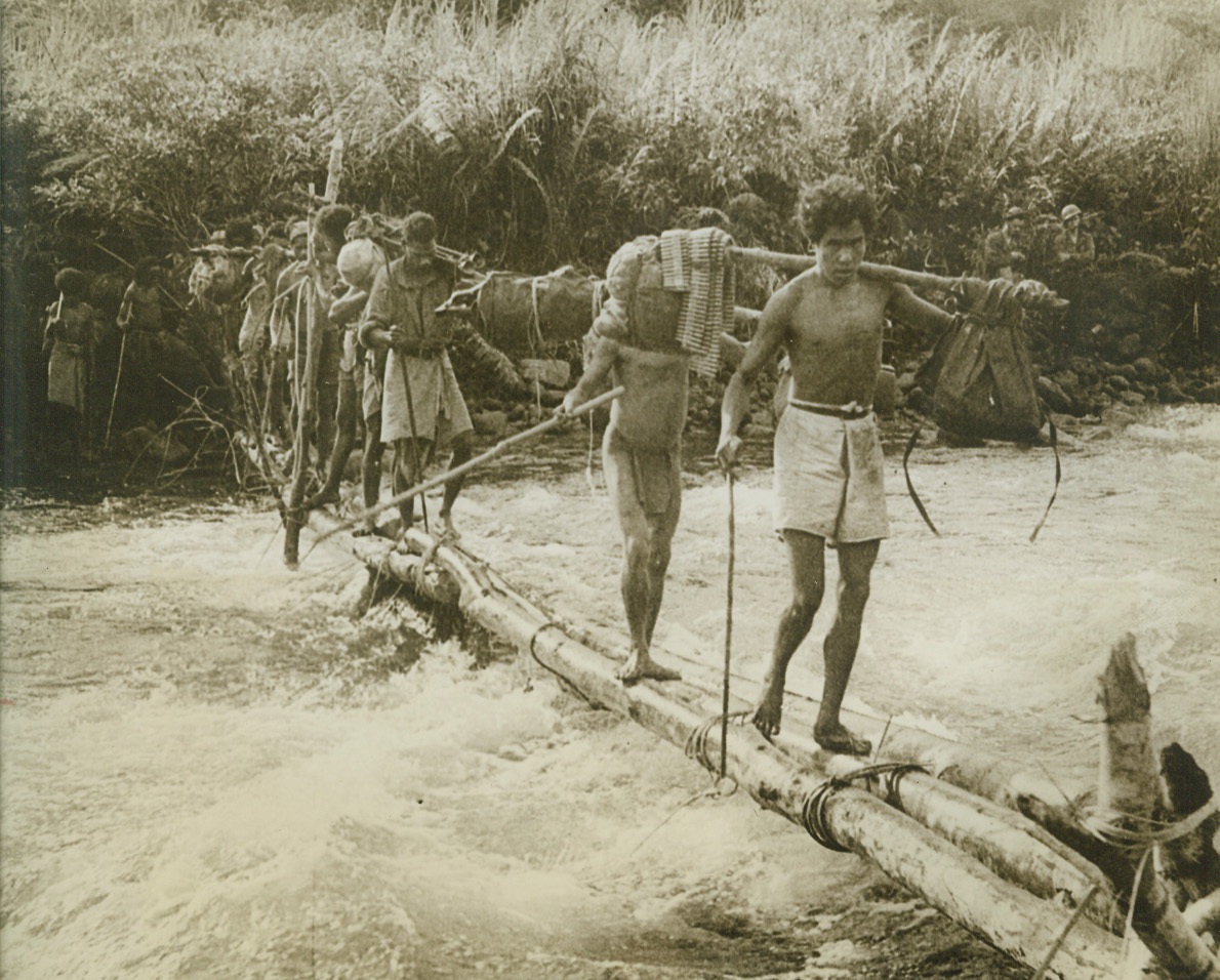 PRECARIOUS CROSSING, 12/19/1942. NEW GUINEA—Sure-footed natives carry equipment and ammunition across a bridge, set up over rushing waters by American troops, in theheart of the New Guinea jungle. After being flown across New Guinea, American and Australian troops had to hike through jungles for six days before they were able to engage the Japs at Buna. Recent reports from Buna indicate heavy fighting in that area, while United Nations bombers carry out raids on enemy installations on Timor Island. Credit: Acme;