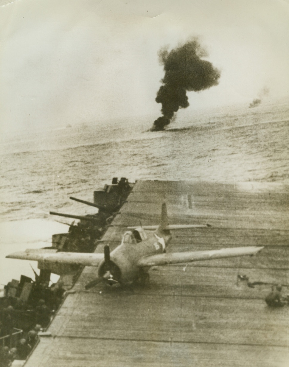 Fighting in the Pacific, 12/23/1942. A U.S. Navy plane came to an abrupt stop on the deck of a zig-zagging aircraft carrier during a battle in the Pacific.  A member of the plane’s crew, or the deck crew, crouches at right as the ship dodges Jap planes.  Two columns of smoke rise from the ocean, possibly showing where planes have crashed during the fight.  Photo from an official Navy film. Credit (Official Navy photo – ACME);