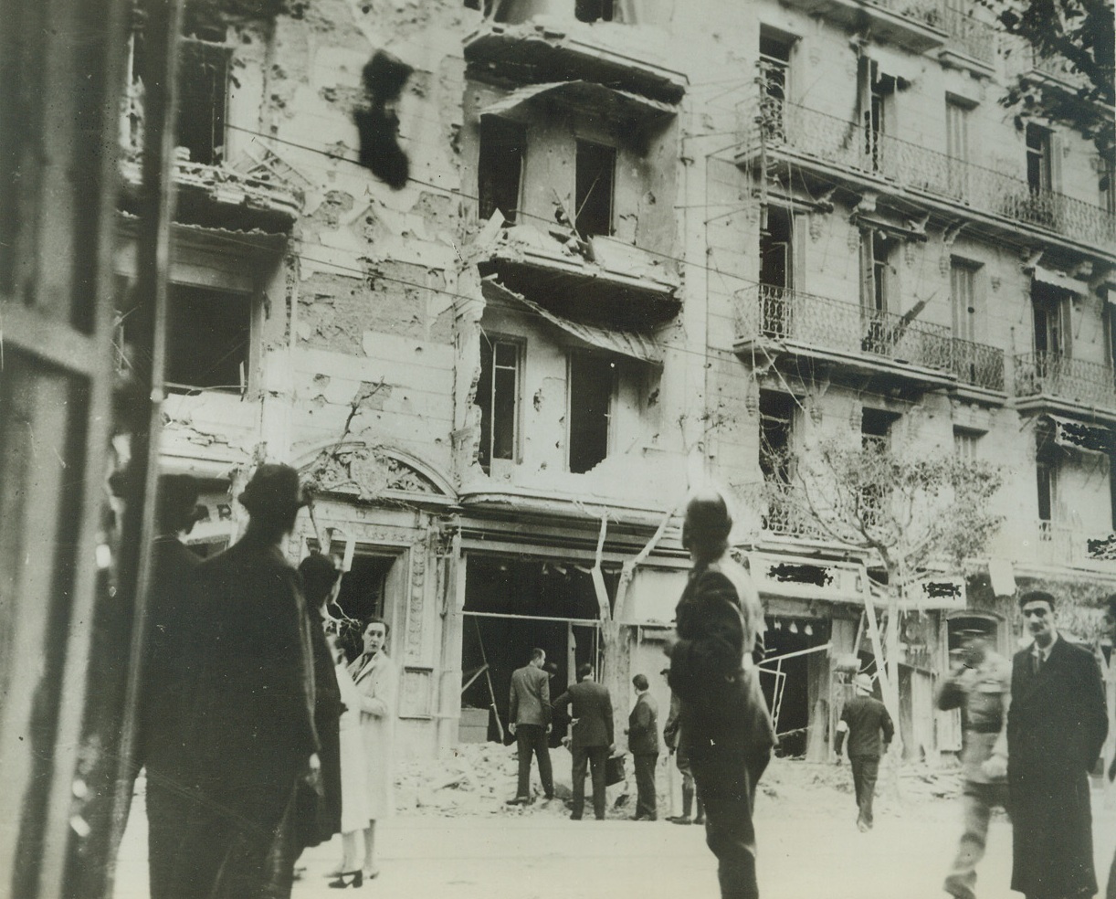 Nazi Raiders Damage Algiers, 12/13/1942. Algiers—Citizens of Algiers curiously inspect the damage done to a building during the first German raid on the territory following the occupation by Allied troops. The man wearing a helmet and arm badge, about to enter the least-damaged building, apparently is a civilian air raid warden. Credit: ACME.;