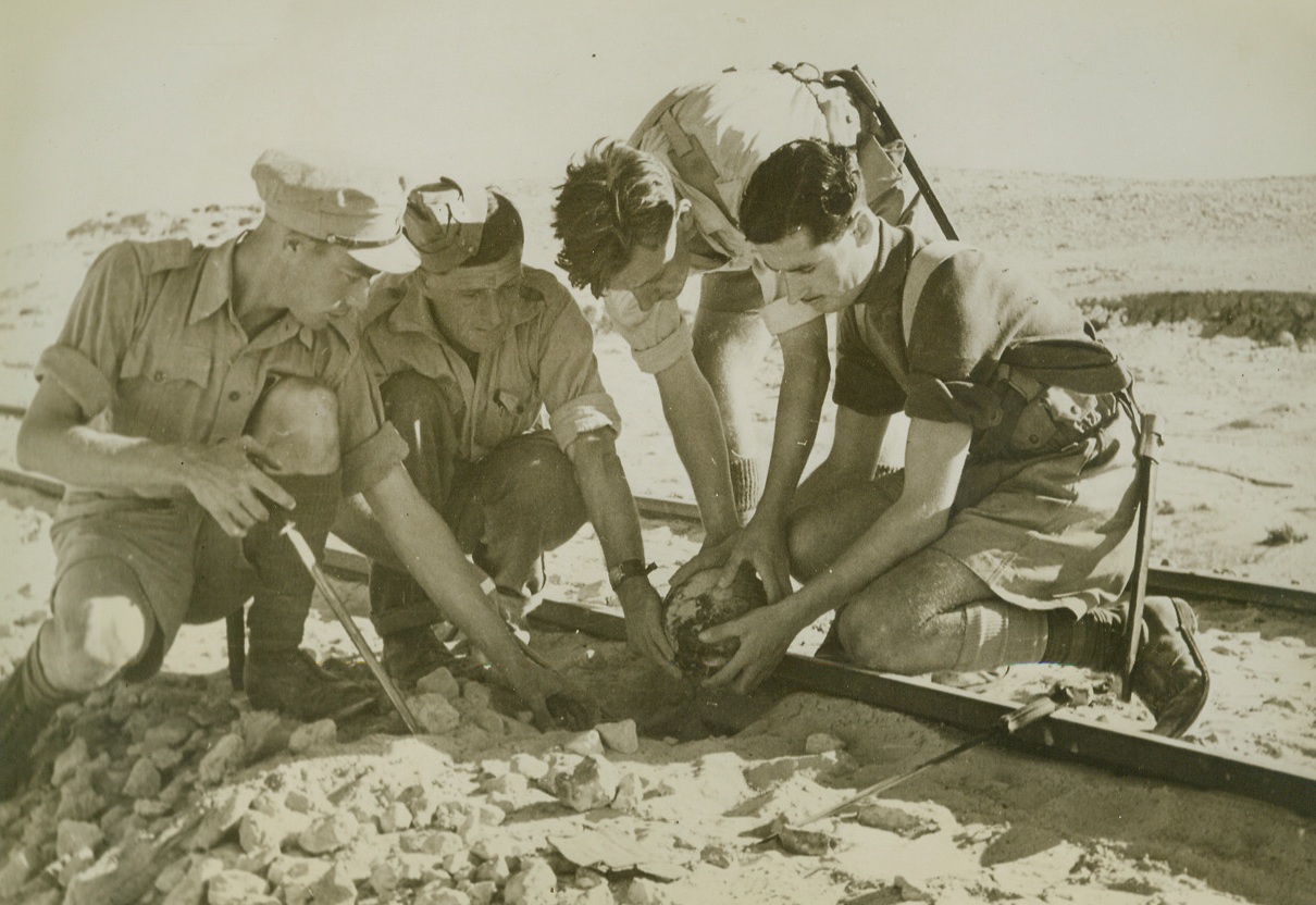 Rebuilding Desert Railroad (#1), 12/9/1942. As Allied forces pursued German Marshal Rommel’s fleeing Afrika Korps toward the west, driving the Axis out of Egypt, British engineers had to rebuild roads and railroads. Every inch of both roads and rail lines had to be searched for mines and booby traps. Here, sappers have located and are removing a mine laid under a rail by the Axis. Note bayonets which were used to probe for mines. Passed by censors.Credit: ACME.;